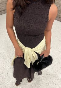 Brun tekstureret ærmeløs kjole med rynket design, bundet gul tørklæde i taljen og en sort pels taske med sølvdetaljer.