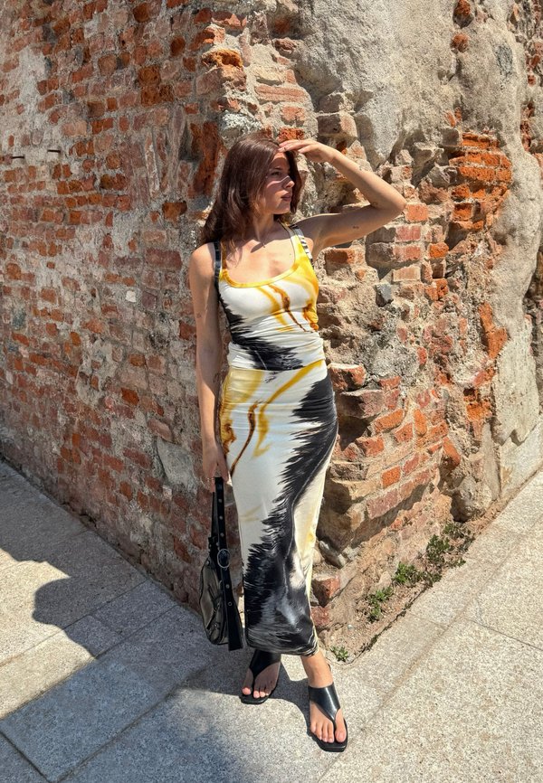 Femme se protégeant les yeux du soleil, portant une robe à motifs jaune, blanche et noire, des sandales noires, et tenant un sac à main noir près d’un mur de briques usé.