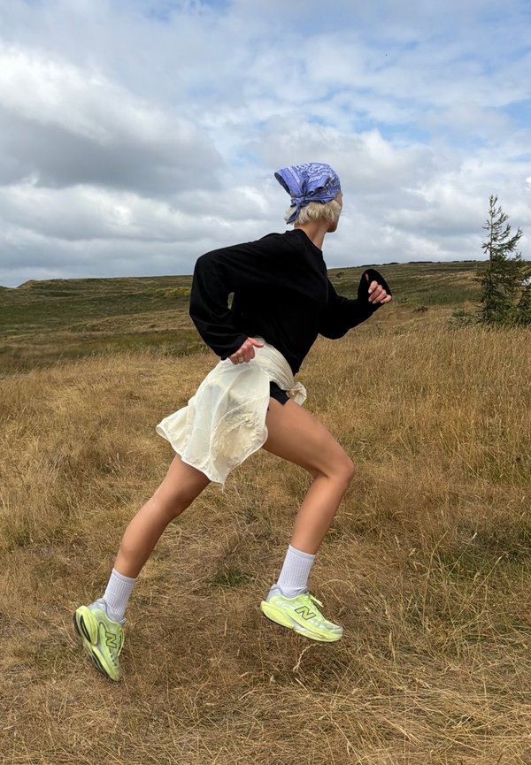 Isik, kellel on peas sinine bandana, must pika varrukaga särk ja neoonrohelised jalanõud, jookseb ülesmäge kuivas rohuses väljas, pilvise taeva all.