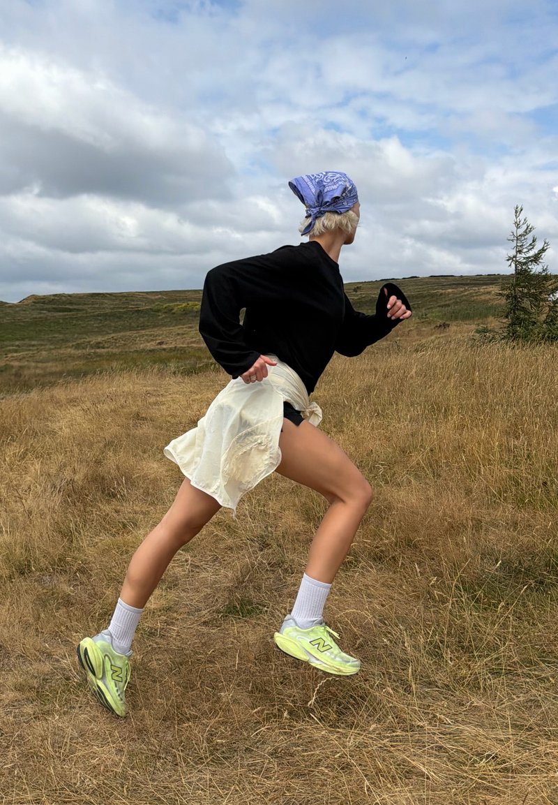 Personne portant un bandana bleu, un haut à manches longues noir et des chaussures vert fluo courant en montée dans un champ herbeux sec sous un ciel nuageux.