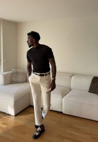 Homme en t-shirt noir et pantalon beige debout dans un salon avec un canapé d'angle blanc, tenant un petit récipient dans sa main droite.