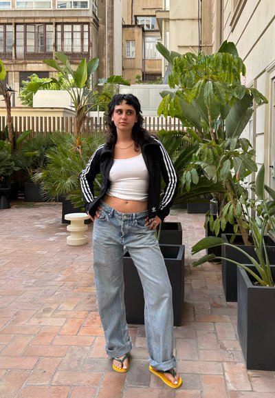 Chaqueta negra de Adidas con cremallera y rayas blancas, top corto blanco, jeans holgados azul claro y chanclas amarillas con correas verdes.