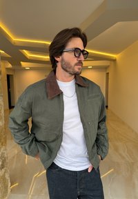 Olívazöld bomber dzseki barna kordbársony gallérral, oldalzsebekkel és bordázott mandzsettákkal, fehér ingre és sötét farmerra viselve.