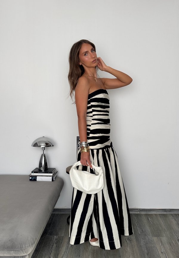 Vrouw draagt een zwart-wit gestreepte strapless jurk, houdt een witte handtas vast en staat naast een stapel boeken en een kleine lamp.
