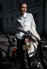 Balta marškiniai su didelėmis kišenėmis, derinami su juodomis kelnėmis. Žalia struktūruota rankinė su auksiniu ženklinimu ir kreminė šalikas, mestas ant pečių.