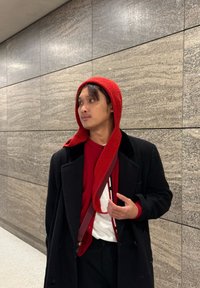 Une capuche rouge en tricot tombe sur un manteau noir, associée à un pull rouge et un pantalon noir. Un mur de pierre lisse et gris en arrière-plan.