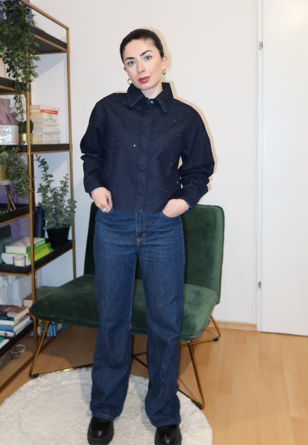 Jachetă din denim închis la culoare, cu închidere cu nasturi și sclipici subtil, asortată cu blugi albastri cu croială dreaptă și cizme negre cu platformă. Scaun verde în fundal.