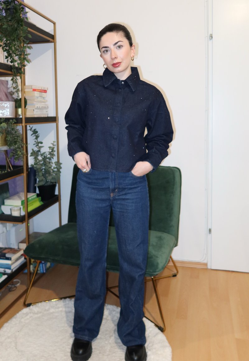 Tume teksajope nööpkinnituse ja kerge sädelusega, kombineeritud siniste sirgete teksapükstega ja mustade platvormsaabastega. Taustal roheline tool.