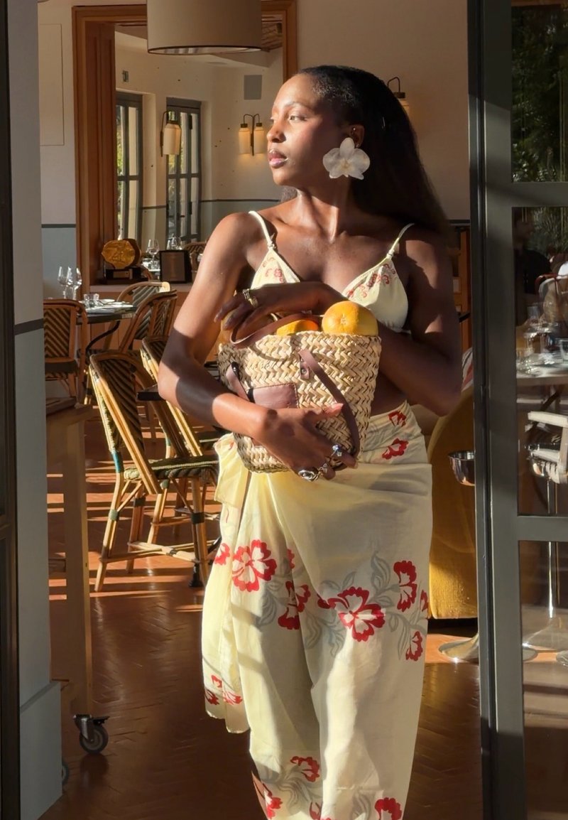 Frau in gelbem Blumenkleid hält einen geflochtenen Korb mit Orangen und steht in einem sonnenbeschienenen Restaurant nahe einer Türöffnung.