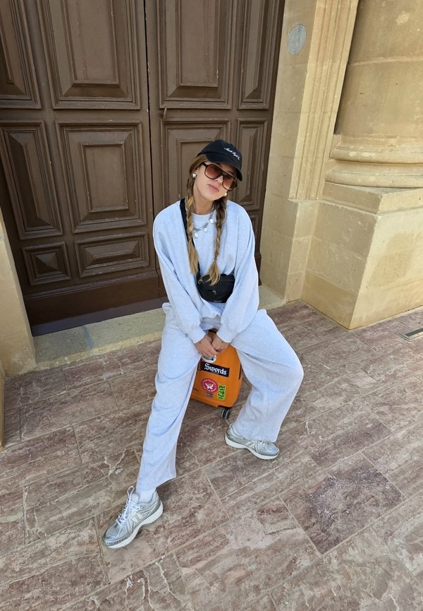 Grauer übergroßer Sweatshirt und Jogginghose mit strukturiertem Finish, kombiniert mit grauen Sneakers. Schwarze Kappe und Sonnenbrille. Ein orangefarbener Koffer.