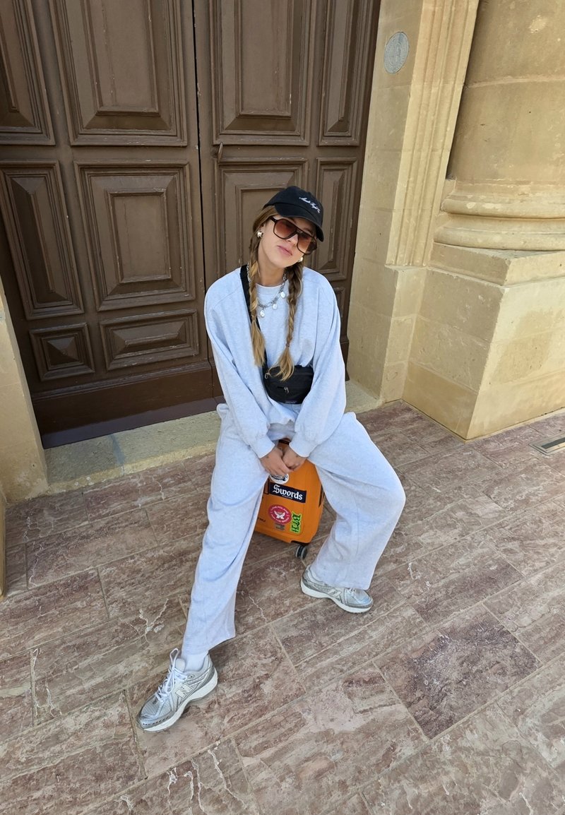 Sweatshirt surdimensionné gris et pantalon de survêtement avec une finition texturée, associés à des baskets grises. Casquette noire et lunettes de soleil. Une valise orange.