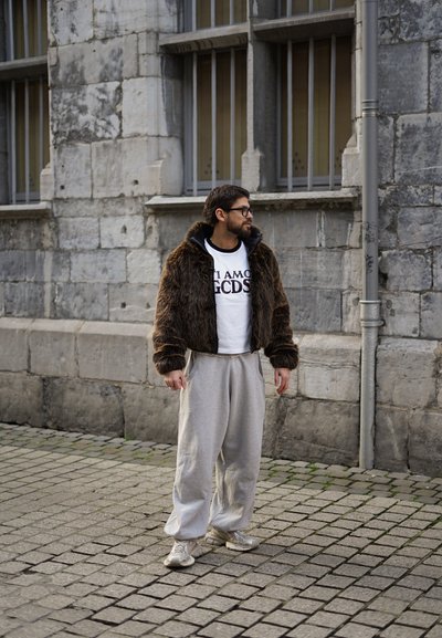 Chaqueta corta de pelo marrón sobre una camiseta blanca con texto negro, pantalones deportivos grises y zapatillas plateadas, de pie en una calle de adoquines.