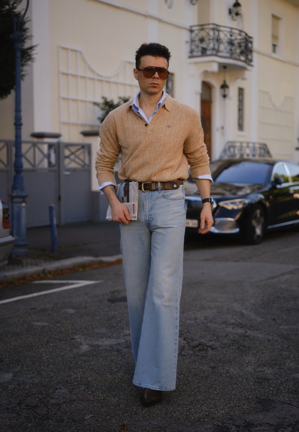 Helles beige Kabelstrickpullover über einem blauen Hemd mit Kragen, kombiniert mit hellblauen Schlagjeans und braunen Schuhen. Große Sonnenbrille und eine Zeitschrift in der Hand.