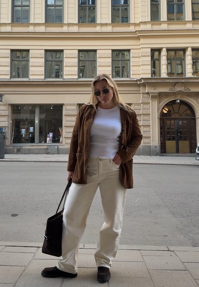 Blazer de ante marrón sobre una camisa blanca, pantalones anchos crema y calzado oscuro, sosteniendo un bolso oscuro contra un fondo de calle de ciudad.