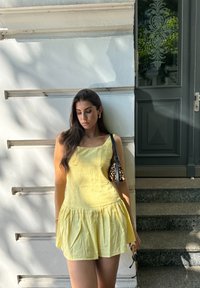 Gelbes Leinenkleid mit quadratischem Ausschnitt und schmeichelndem Rock. Eine kleine Handtasche im Leopardenmuster wird über der Schulter getragen. Steinige Stufen im Hintergrund.