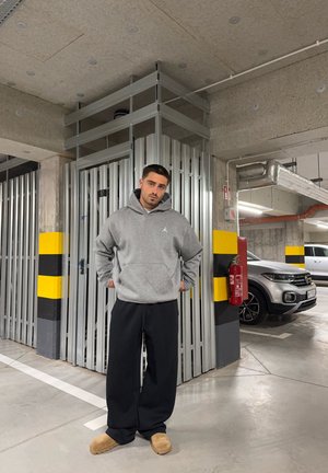 Sudadera gris con bolsillo frontal y logo, pantalones holgados negros y pantuflas color marrón, situados en un garaje con accesorios metálicos.