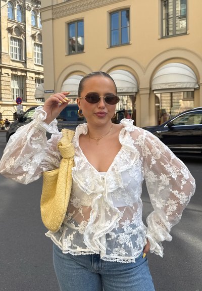 Blusa de encaje blanca con bordes rizados y mangas largas, combinada con jeans de color azul claro. Sosteniendo un bolso de mimbre amarillo y usando gafas de sol.