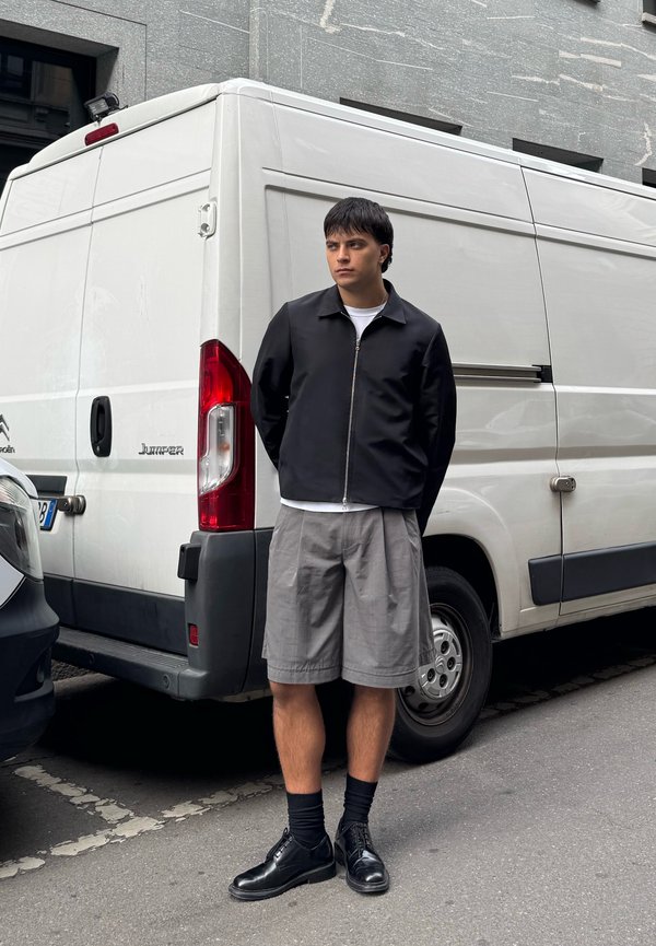 Giacca nera con zip, T-shirt bianca, pantaloni corti grigi, calzini neri e scarpe. In piedi accanto a un furgone bianco in una strada cittadina con uno sfondo di edifici grigi.
