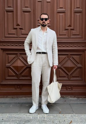 Traje de lino de color claro con un acabado texturizado, combinado con una camisa blanca y zapatillas blancas. Sostiene un bolso tote beige. Fondo de madera oscura.