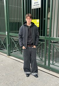 Veste bomber noire sur un sweat à capuche noir, associée à un jean noir ample avec les ourlets pliés. Portée avec des chaussures noires, debout devant une porte verte.