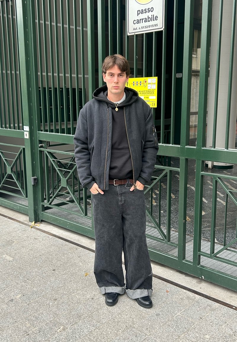 Veste bomber noire sur un sweat à capuche noir, associée à un jean noir ample avec les ourlets pliés. Portée avec des chaussures noires, debout devant une porte verte.