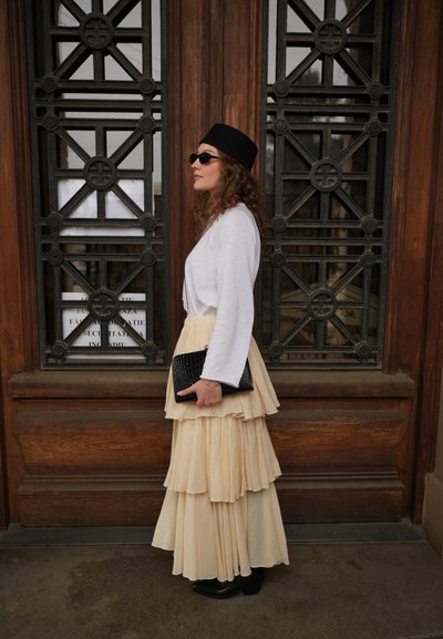 Mujer con falda beige de capas, camiseta blanca de manga larga, sombrero negro y gafas de sol sostiene un bolso negro de mano junto a una puerta de madera con rejas de hierro.