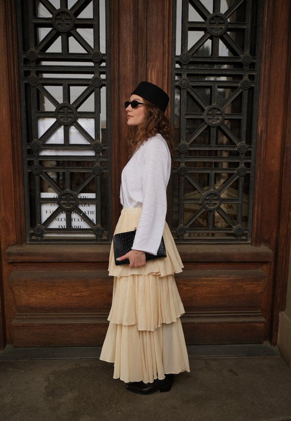 Kvinde iført lagdelt beige nederdel, hvid langærmet top, sort hat og solbriller, holder en sort clutch og står ved en trædør med jerngitre.