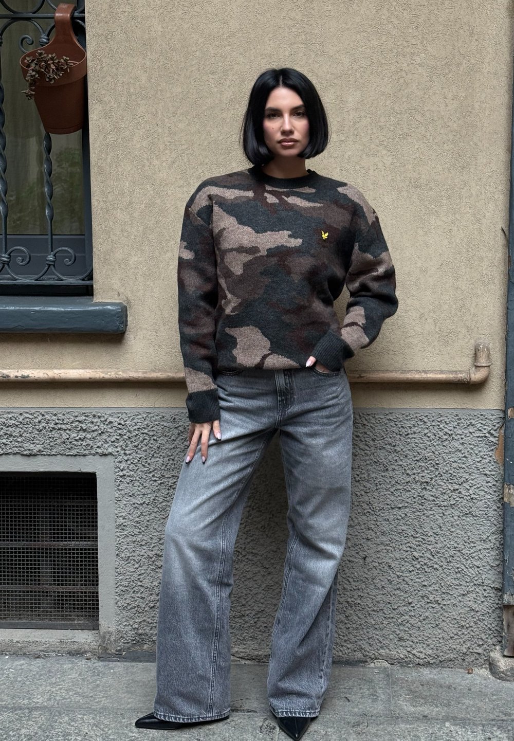 Sweter w camo w ciemnych tonach z małym żółtym logo, zestawiony z jasnoszarymi szerokimi spodniami i czarnymi, czubkowatymi butami.