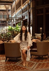 Robe légère et fluide avec des manches surdimensionnées dans un motif crème. Chaussures beige et un petit sac à main pailleté. Environnement intérieur avec des plantes.