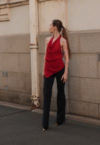 Top halter rosso drappeggiato con un design fluido abbinato a pantaloni neri attillati. L'outfit presenta tacchi a spillo neri con accenti dorati.