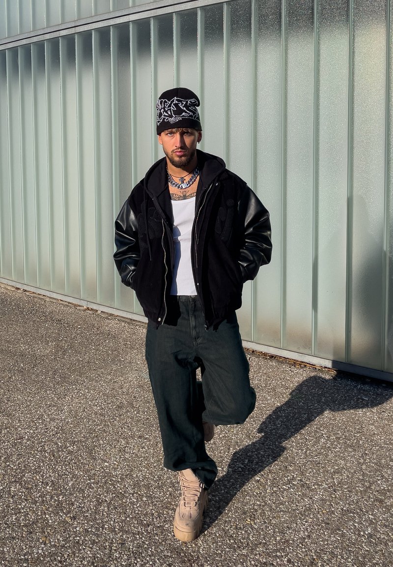 Homme portant un bonnet noir et une veste, débardeur blanc, pantalons foncés et bottes beige, debout les mains dans les poches contre un mur en verre translucide.