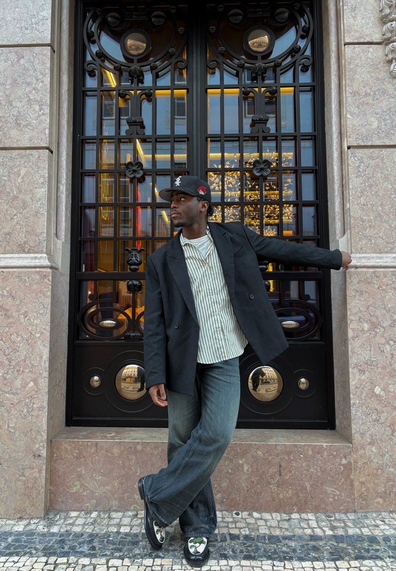 Homme en blazer noir, chemise rayée, casquette de baseball et jean, appuyé de côté contre une grille décorative en métal noir avec des reflets et des murs en pierre.