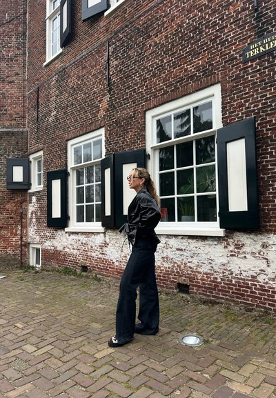 Chaqueta de cuero negra con cintura ajustable, pantalones anchos oscuros y zapatos estampados, contra un muro de ladrillo rojo con persianas negras.