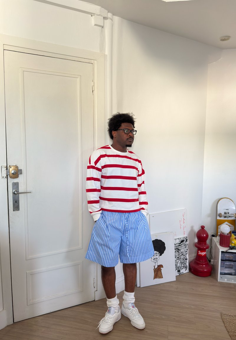 Ung mann med briller står innendørs iført en rød og hvitstripet skjorte, blå og hvitstripete shorts, hvite sokker og hvite joggesko.