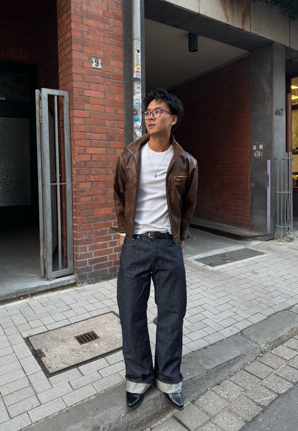 Joven con gafas, vestido con una chaqueta de cuero marrón, camisa blanca y vaqueros anchos y oscuros, de pie en una calle empedrada junto a un edificio de ladrillo.
