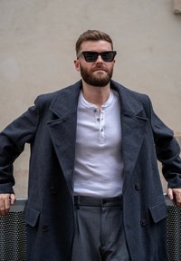 Lungo cappotto di lana grigio scuro con ampi risvolti, camicia henley bianca e pantaloni grigi. Persona che indossa occhiali da sole neri, in posa contro un muro.