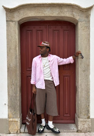 Hombre con camisa rosa, gorra marrón y pantalones cortos a cuadros frente a unas puertas dobles rojas, sosteniendo gafas de sol y un bolso tote marrón.