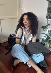 Mladá žena s kudrnatými vlasy sedí na dřevěné podlaze, má na sobě béžovou košili, světle modré džíny a černé boty, drží tmavě šedý kabát, poblíž rostlin a dveří.