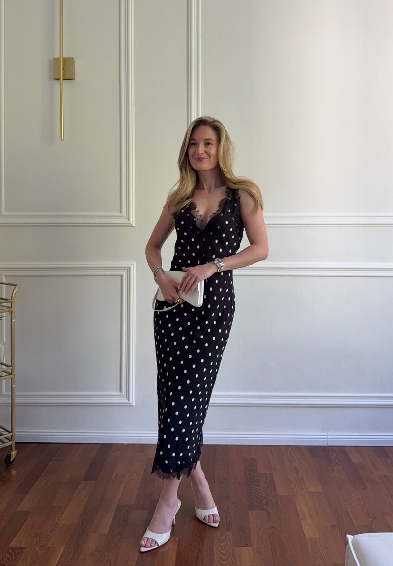 Vrouw staand binnenshuis, gekleed in een zwarte polkadotjurk met kanten afwerking, witte sandalen met hak, houdt een witte clutch vast en glimlacht.