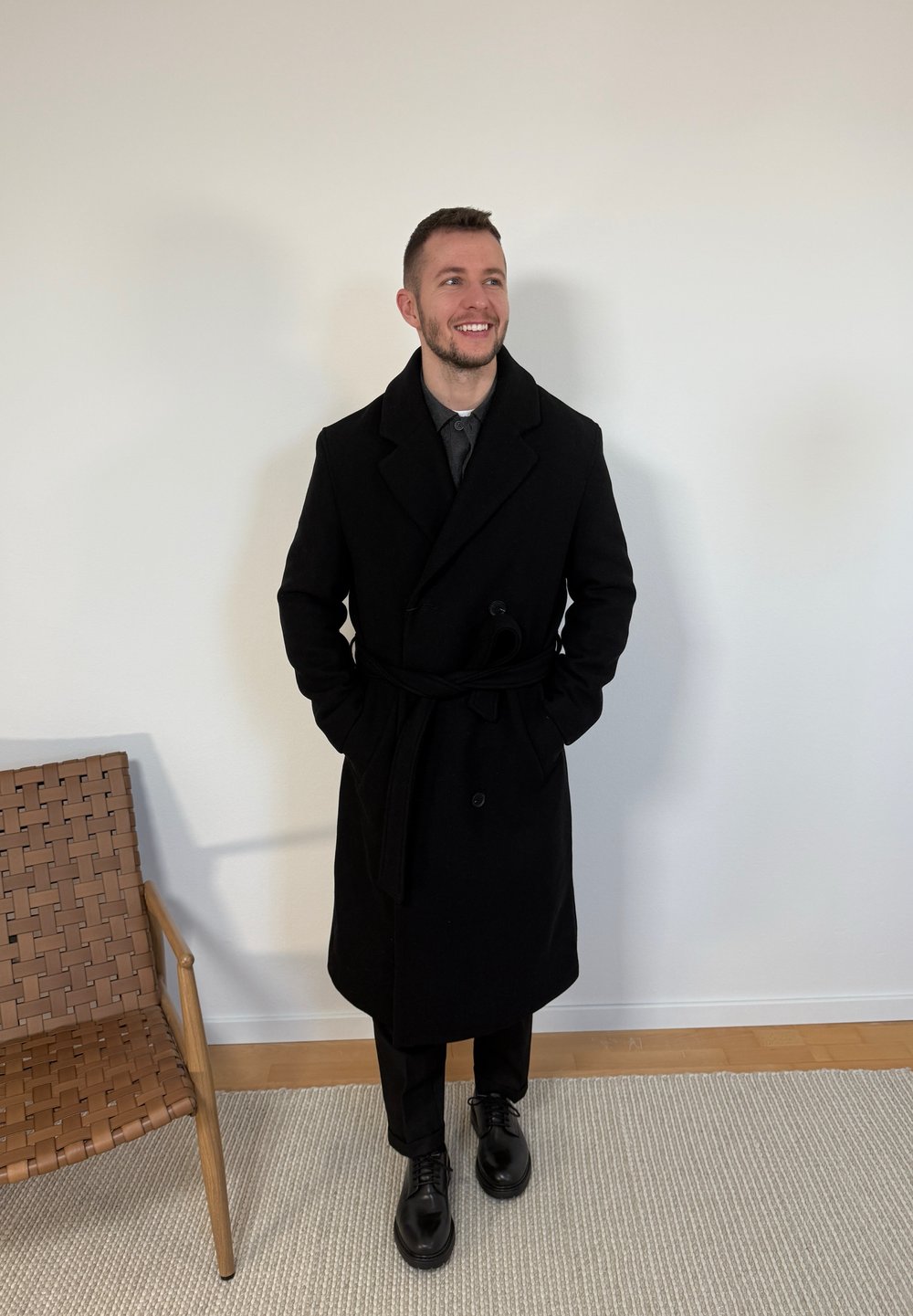 Άνδρας που χαμογελά, φορώντας ένα μακρύ μαύρο παλτό, μαύρο παντελόνι και μαύρα παπούτσια, στέκεται μέσα σε ένα χώρο δίπλα σε μια υφαντή ξύλινη καρέκλα πάνω σε ένα χαλί.