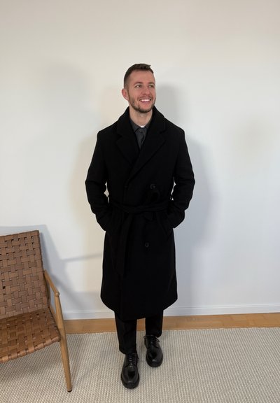 Hombre sonriente, vestido con un abrigo largo negro, pantalones negros y zapatos negros, de pie en el interior junto a una silla de madera tejida sobre una alfombra.