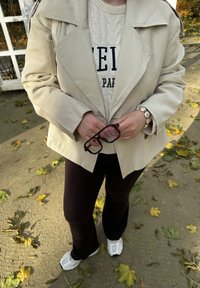 Beige trenssitakki leveillä kauluksilla vaalean kermanvärisen, mustilla kirjaimilla koristellun palmikkoneuleen päällä. Yhdessä tummien kehysten silmälasien kanssa, päällä tummat housut.