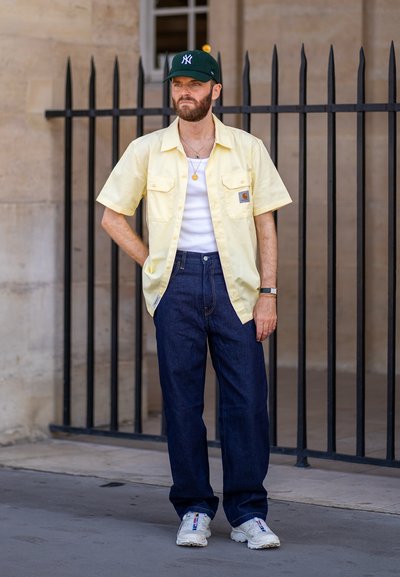 Camisa de manga corta amarillo claro sobre una camiseta blanca; jeans rectos azul oscuro; zapatillas deportivas blancas; gorra verde con logo; collares en capas.