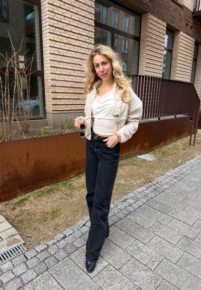 Mujer con cabello largo y rubio, vistiendo chaqueta blanca, camiseta blanca y pantalones negros, sosteniendo gafas de sol, de pie en una acera pavimentada cerca de un edificio de ladrillo.