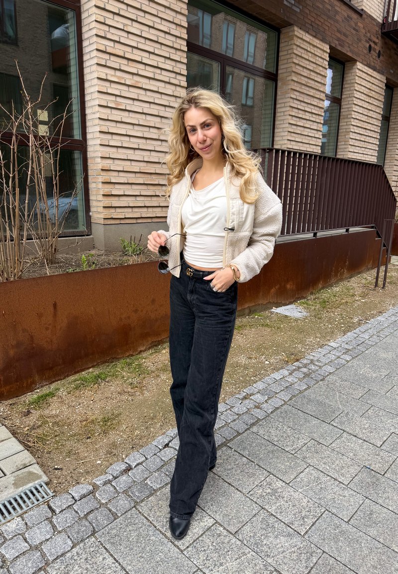 Kvinde med langt blondt hår iført hvid jakke, hvid top, sorte bukser, holder solbriller, stående på en belagt fortov nær en murstensbygning.