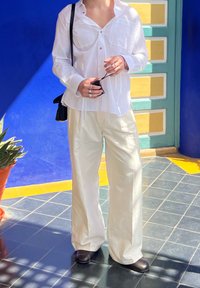 Camicia bianca a maniche lunghe con due tasche sul petto, pantaloni bianchi larghi e scarpe scure. Borsa a tracolla nera e anelli sulle mani.