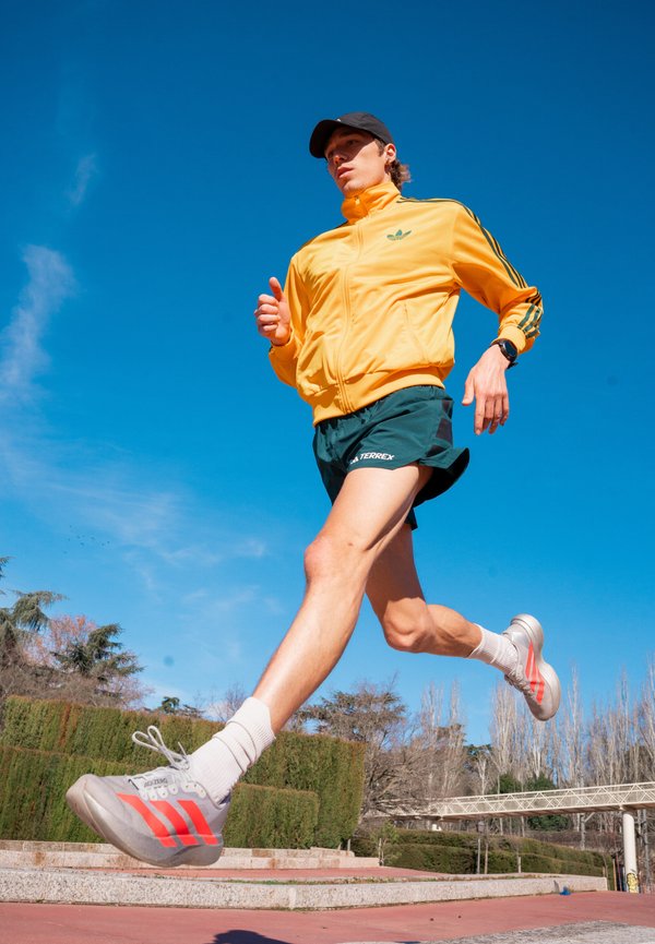 Gul zip-up jakke med grønne striper, grønne shorts og grå joggesko med røde detaljer. Personen er i midtgang på en bane.