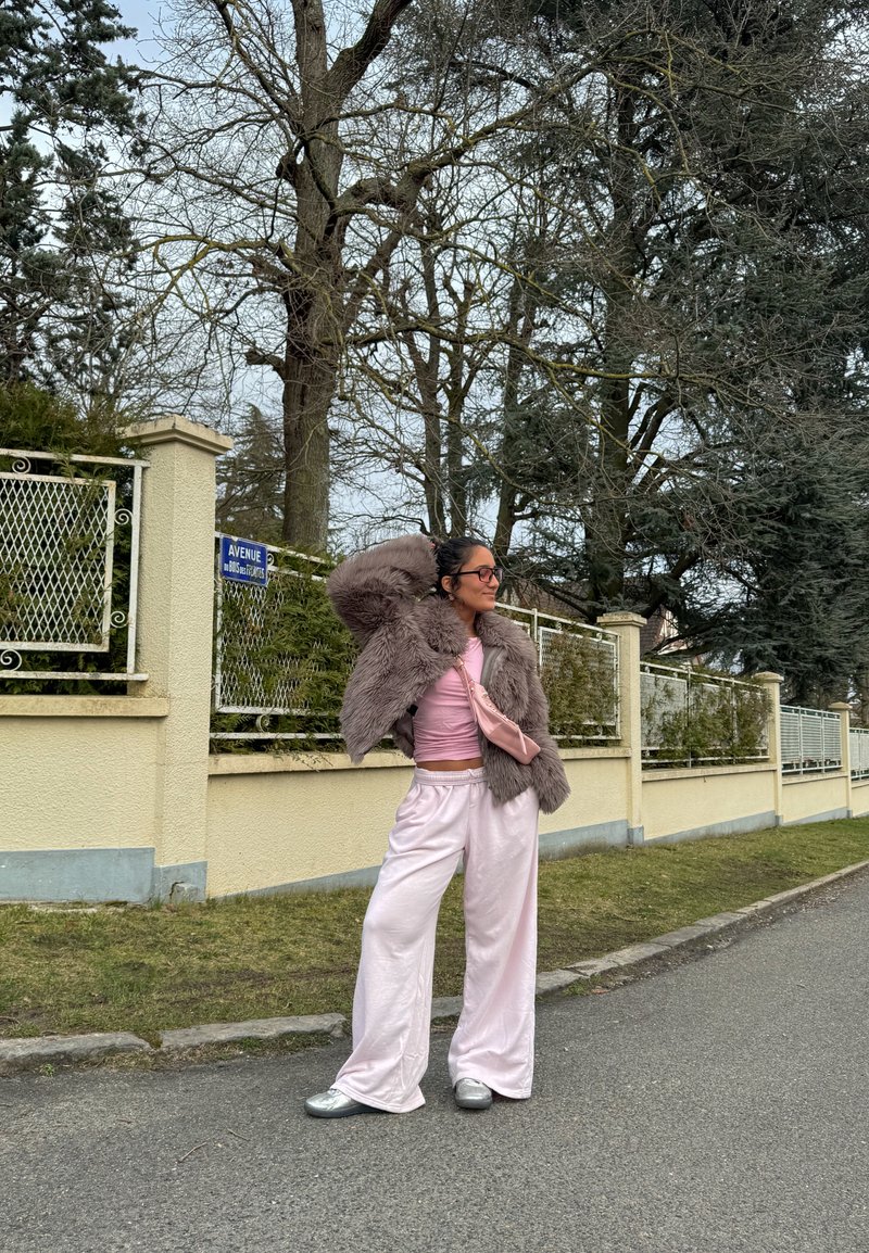 Personne portant un haut rose, un pantalon large, une veste en fourrure grise et un sac rose, debout dans la rue près d'une clôture beige avec des arbres nus en arrière-plan.