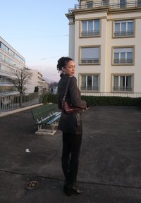 Jupe en cuir foncé, pull gris côtelé, bottes noires montant jusqu'aux genoux, et sac bandoulière bordeaux. Cadre extérieur épuré avec des bâtiments modernes.