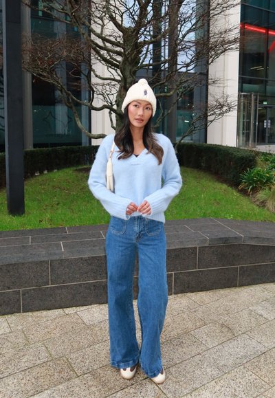 Mujer que lleva un gorro de punto blanco, suéter de felpa azul claro, vaqueros de pierna ancha y zapatos blancos, de pie en un camino pavimentado cerca de un árbol sin hojas y edificios urbanos.
